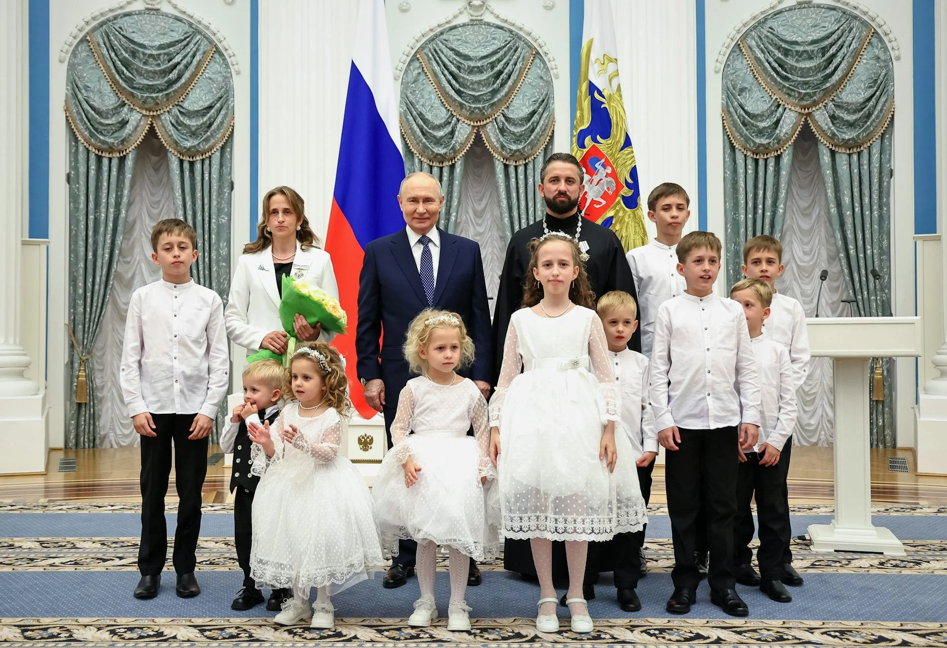 Two men and a woman stand with 10 children in white outfits in an ornately decorated room.