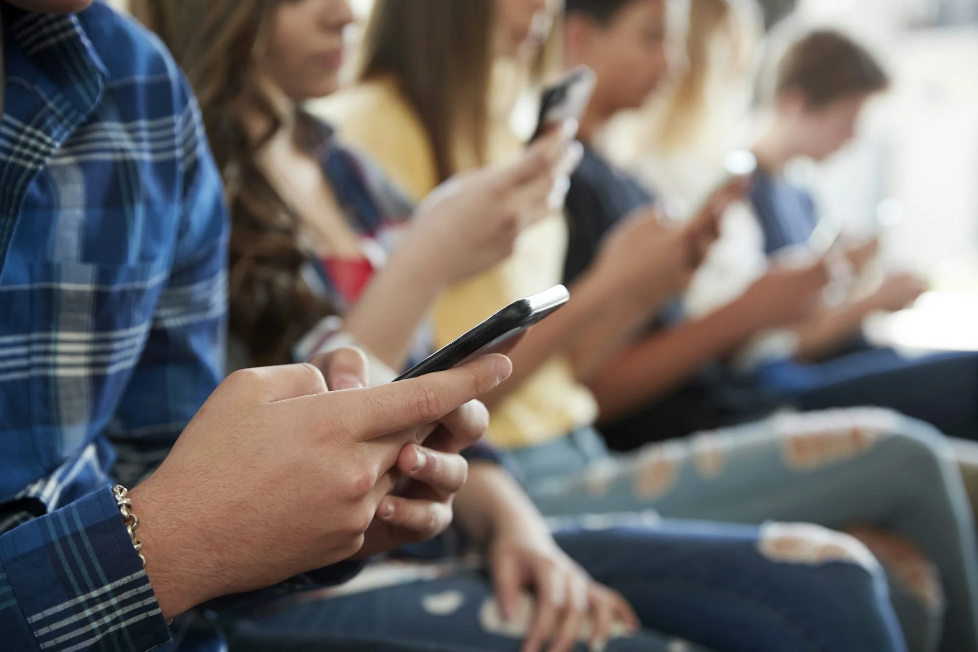 A row of teens sit next to each other while each looksat a cellphone screen