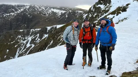 Matty Belcher | Brandan Smith Three men in winter coats and backpacks stand on top of a high snow covered rocky ridge with a mountain peak behind them.