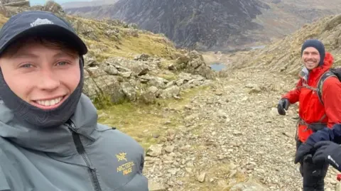 Matty Belcher | Brandan Smith A man with a dark blue baseball cap and grey windbreaker smiles at the camera. To his left stands a man in a red jacket and blue hood, with a large hiking backpack. Behind them is a rocky mountain and blue lake in the valley below.