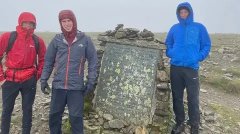 Matty Belcher | Brandan Smith Three young men in red, grey and blue winter jackets stand at the top of a cloud covered mountain top with their hoods pulled tightly around their faces. 