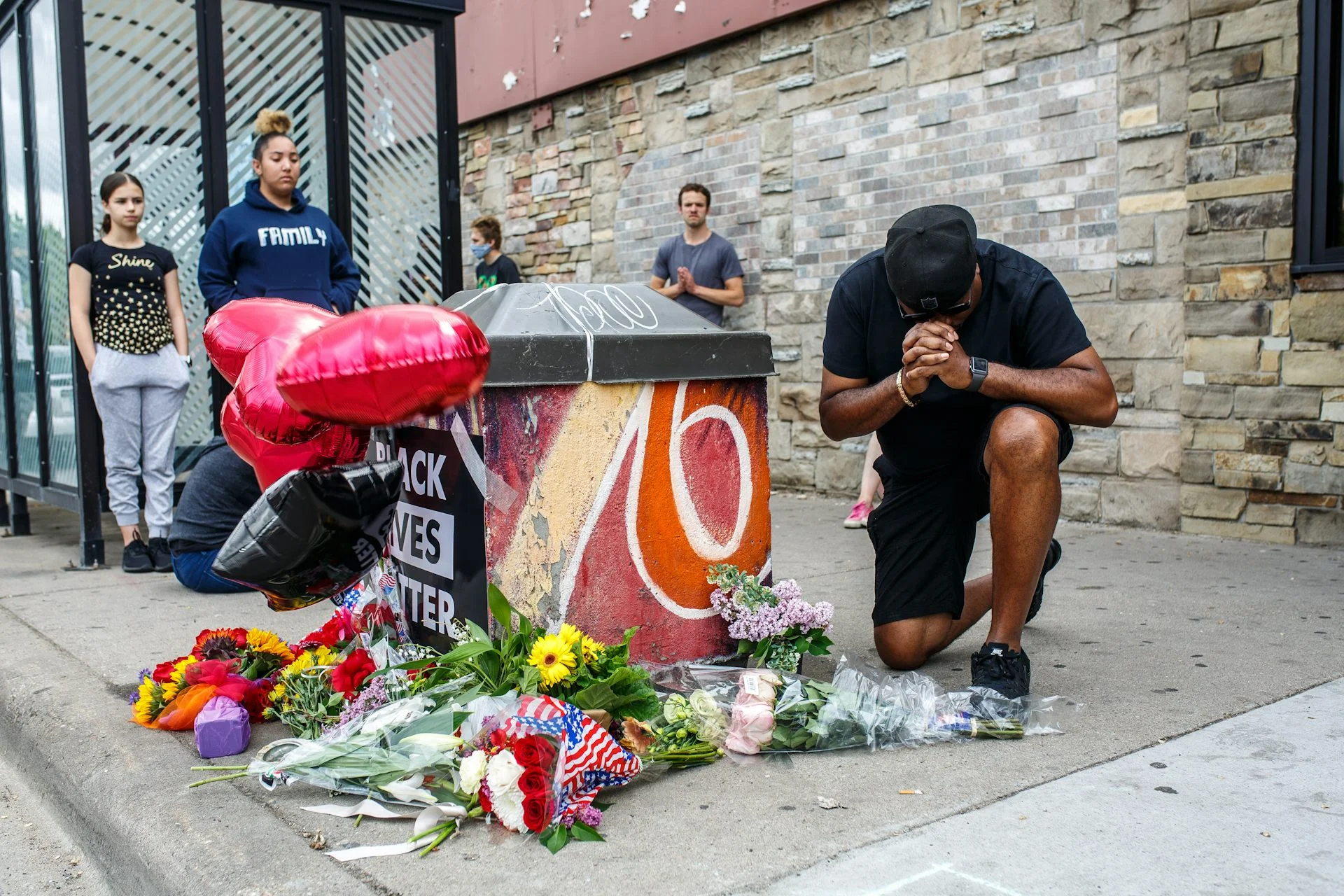 A man kneels in front of a makeshift memorial next to a bus stop on a city street.